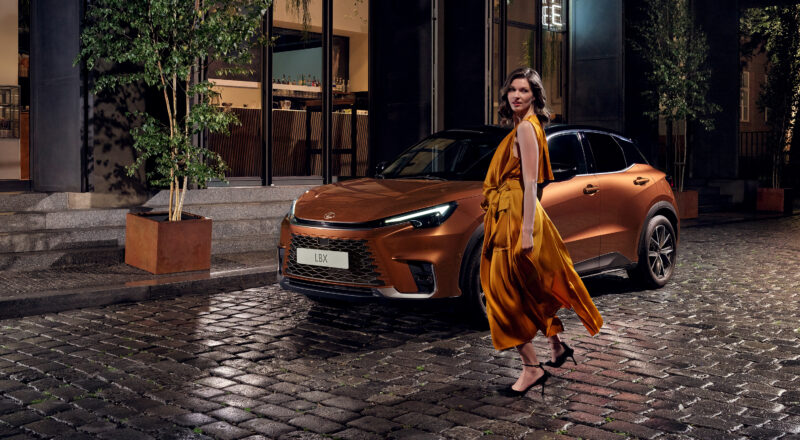 Lexus LBX color cobre estacionado en calle urbana de noche con modelo caminando frente al vehículo.