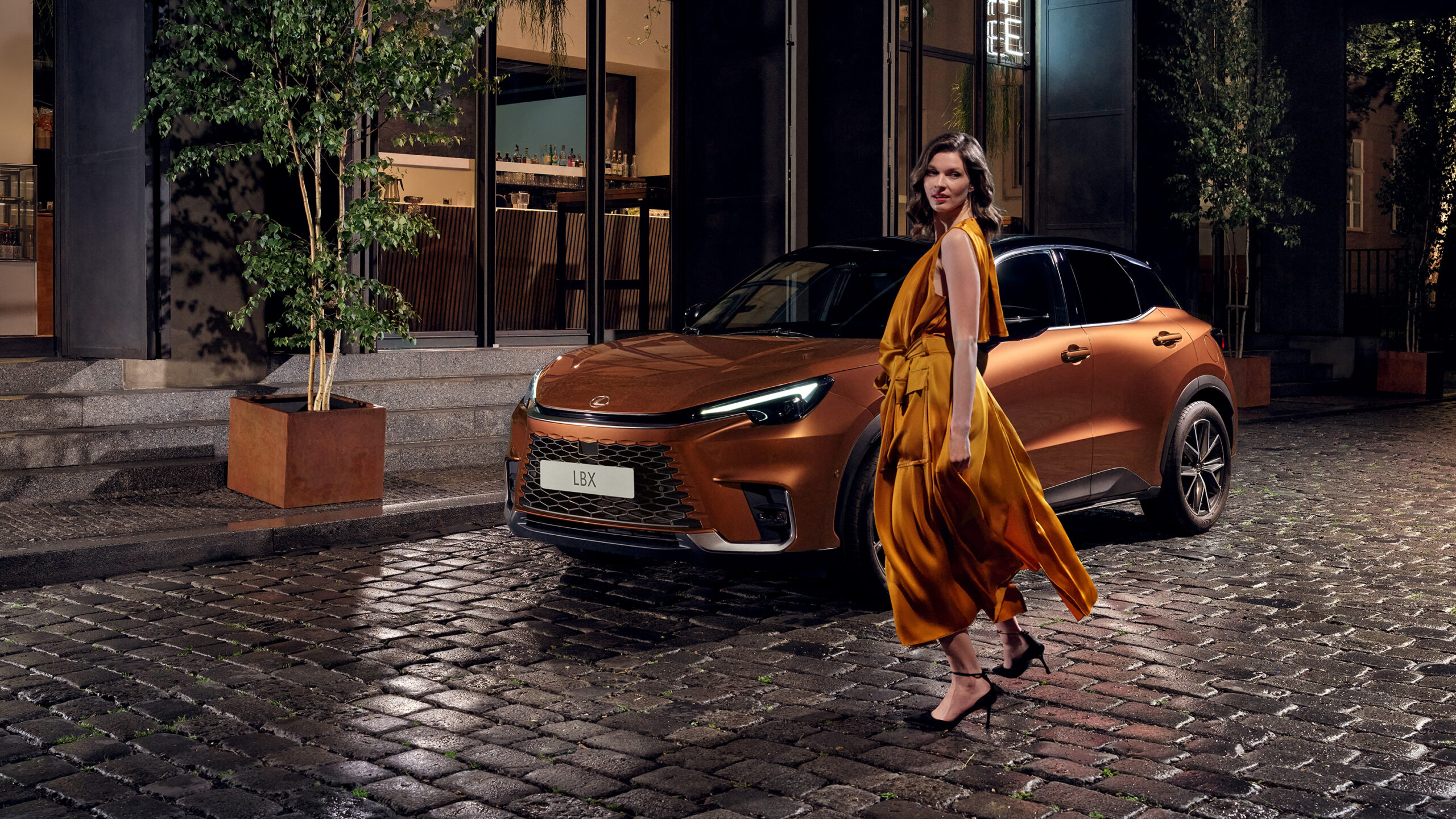 Lexus LBX color cobre estacionado en calle urbana de noche con modelo caminando frente al vehículo.