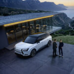 Lexus TZ blanco junto a una pareja frente a una vivienda panorámica al atardecer.