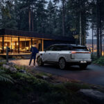Mujer cargando un Lexus TZ blanco frente a una casa moderna en un bosque al anochecer.